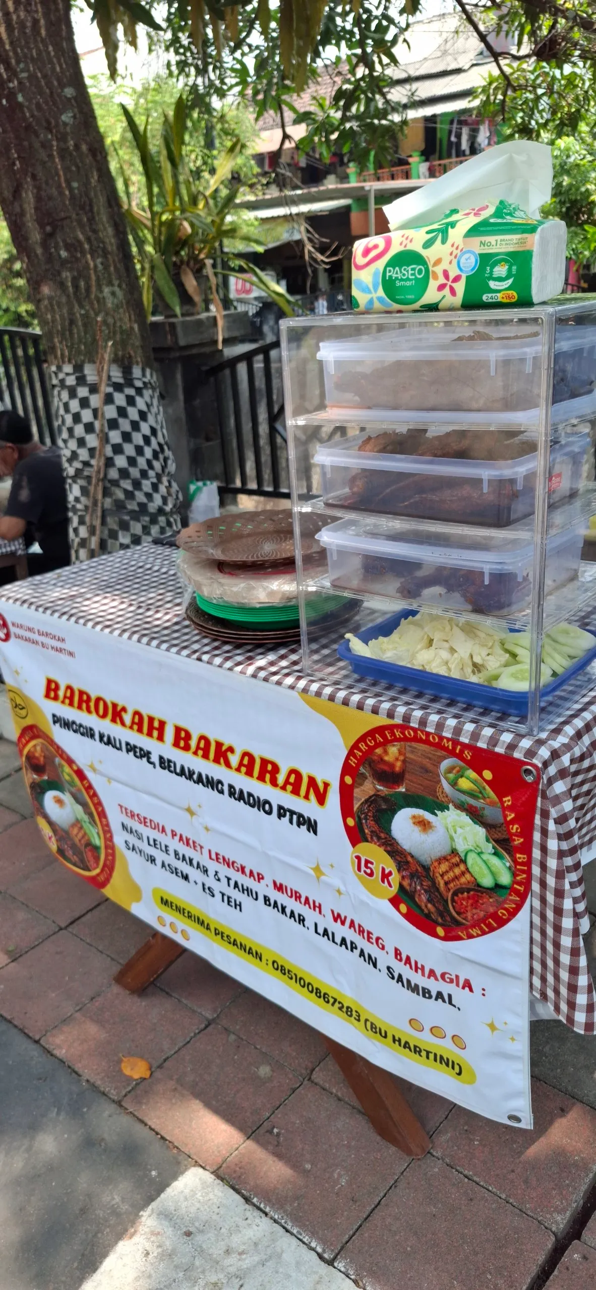 Lapak Barokah Bakaran di bawah pohon dengan spanduk alamat Pinggir Kali Pepe dan nomor pesanan Bu Hartini.