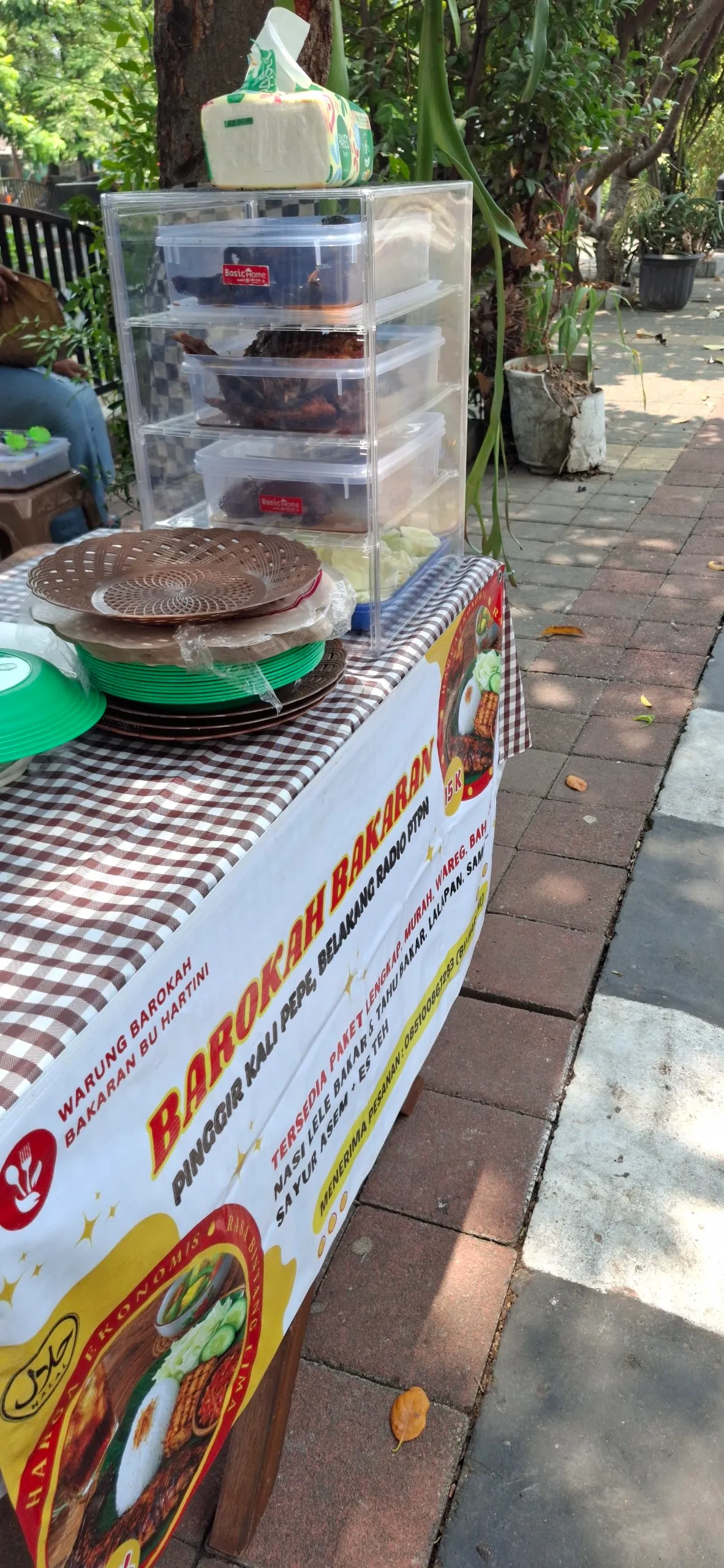 Meja lapak bertaplak kotak-kotak di trotoar, dengan etalase lauk dan tumpukan piring.