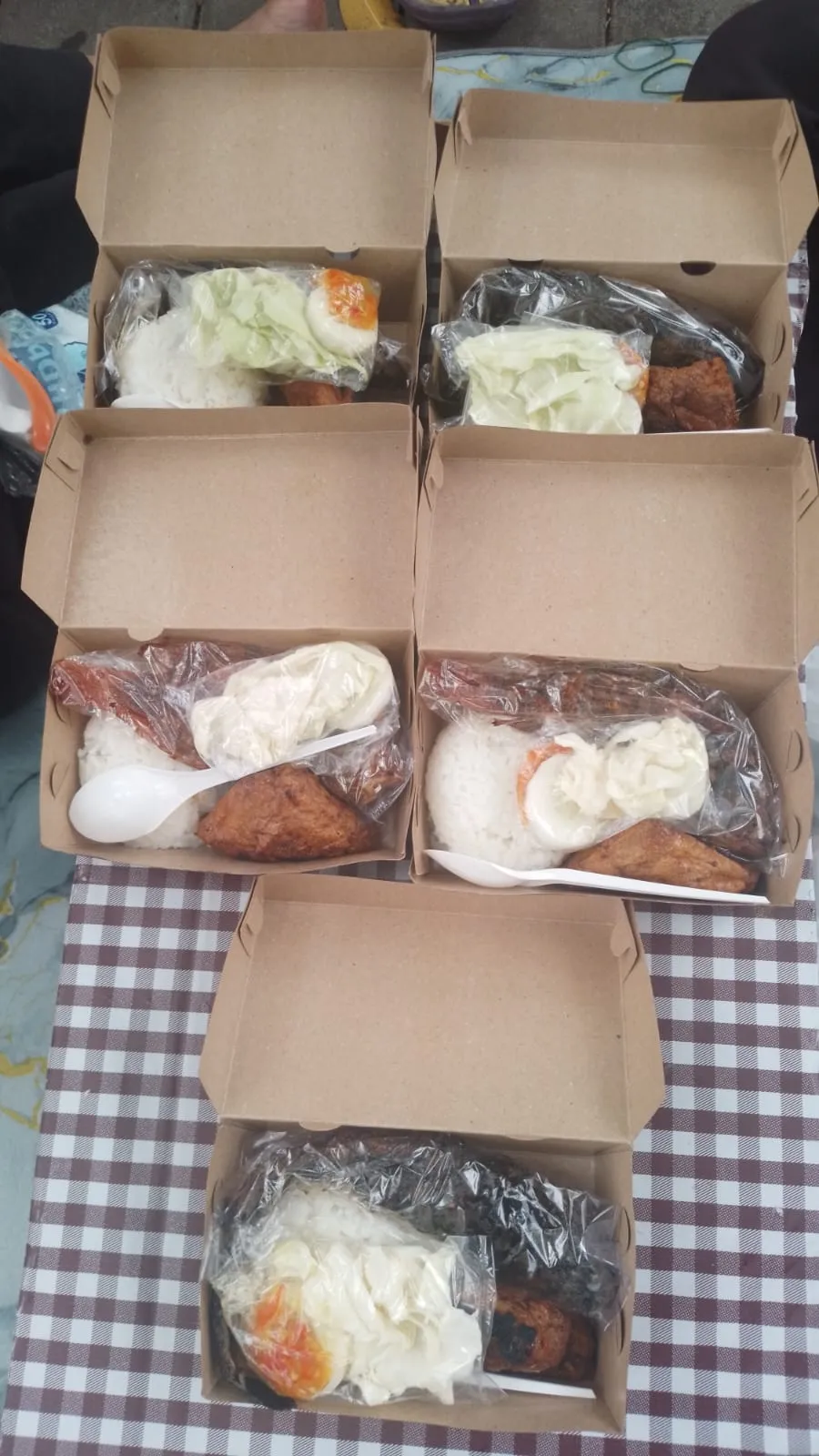 Enam kotak berisi nasi, lauk bakar, telur, dan lalapan di atas taplak kotak-kotak.