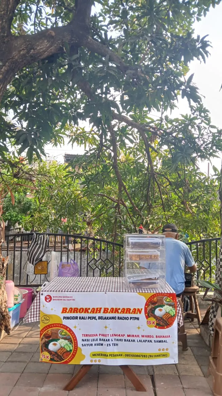 Warung Barokah Bakaran di bawah pohon rindang, dengan spanduk menu dan etalase makanan di Pinggir Kali Pepe.